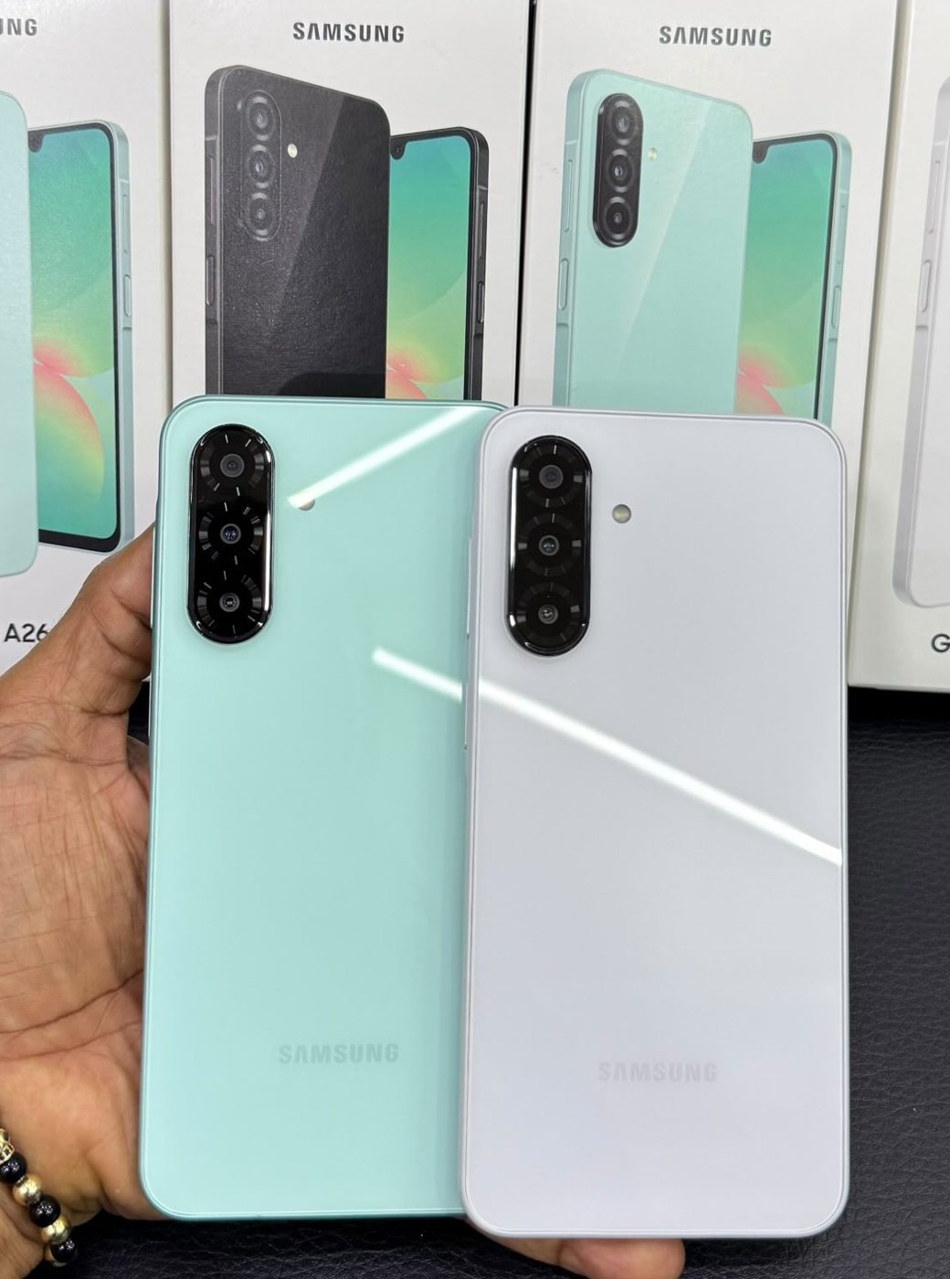 Dos Samsung Galaxy A26, uno verde menta y uno blanco, sostenidos frente a las cajas de la tienda